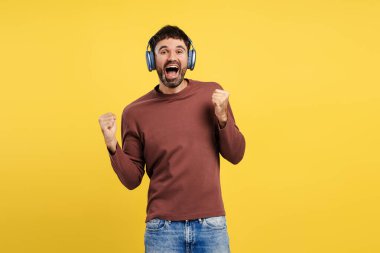 Heyecanlı, orta yaşlı adam kulaklıkla müzik dinliyor, kazanan el kol hareketi yapıyor, sarı arka planda izole edilmiş kameraya bakıyor. Zafer konsepti