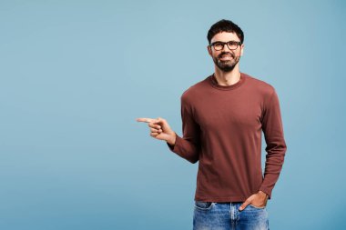 Çekici sakallı, gözlüklü İspanyol bir adamın portresi. Mavi arka planda tek başına duran, cebinde fotokopi çeken elleri işaret ediyor. Alışveriş konsepti