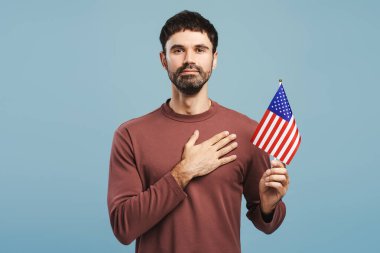 Gülümseyen, mutlu bir Amerikan vatandaşı, elinde Amerikan bayrağı tutan bir vatansever, elini şömineye uzatmış, mavi arka planda izole edilmiş kameraya bakıyor. ABD oylaması, bağımsızlık günü