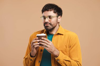 Gözlük takan, aromalı kahve kokan, kahve bardağı tutan, bej arka planda izole edilmiş mutlu bir adamın yakın plan portresi. Kavram kır
