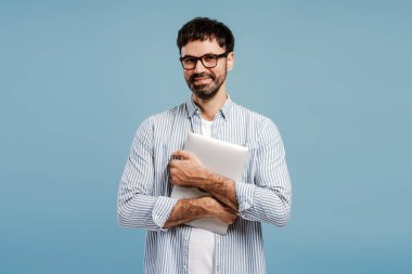 Pozitif, güler yüzlü, şık kıyafetler ve gözlük takan, dizüstü bilgisayarı olan, kameraya bakan, mavi arka planda izole bir şekilde poz veren bir adam. Başarılı programcı. Uzman. Çevrimiçi teknoloji kavramı