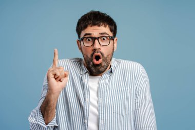 Heyecanlı, şok olmuş sakallı adam parmağını kaldırıyor, şık gözlükler takıyor ve ağzı açık mavi arka planda izole edilmiş kameraya bakıyor. Beyin fırtınası konsepti