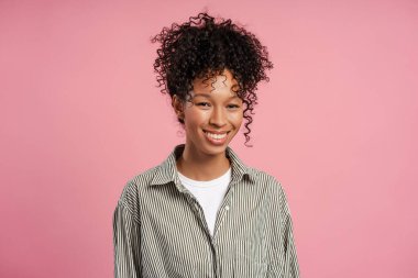 Güzel, gülümseyen Arfican Amerikalı kadın, gömlek giyiyor, kameraya bakıyor, pembe arka planda tek başına duruyor. Halk konsepti 