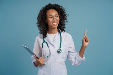 Beyaz önlüklü gülümseyen Afrikalı Amerikalı kadın doktor mavi arka planda izole edilmiş fotokopi alanını işaret ederek başka tarafa bakıyor. Tıp kavramı