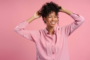 Gülümseyen, çekici Afro-Amerikan kadın gülümseyen ve saçlarına dokunan pembe arka planda tek başına duran kameraya bakıyor. Reklam kavramı 