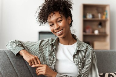 Mutlu, kendine güvenen bir kadın evdeki rahat kanepeye oturmuş kameraya bakıyor. Beyaz dişli, şık saçlı, gülümseyen Afrikalı Amerikalı kadın portresi. Doğal güzellik, sağlıklı yaşam tarzı konsepti