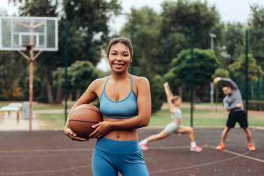 Gülümseyen, mutlu, Afro-Amerikan kadın spor giyimli, kameraya bakarak antrenmandan sonra basketbol oynuyor. Açık havada poz veren pozitif bir sporcu. Etkin yaşam tarzı kavramı