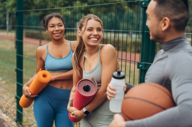 Mutlu, pozitif, çok uluslu kadınlar ve top tutan erkek, yoga minderi ve şişe eğitimden sonra birlikte konuşuyorlar. Dışarıda gülen arkadaşlar. İletişim, spor, motivasyon kavramı