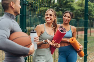 Bir grup pozitif kadın ve erkek top tutuyorlar, yoga minderi ve şişe eğitimden sonra birlikte konuşuyorlar. Dışarıda gülen arkadaşlar. İletişim, spor, motivasyon kavramı