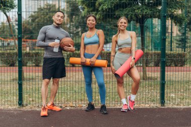Gülümseyen bir grup, çok uluslu erkek ve kadın ellerinde spor paspası şişeyle kameraya bakıyor, dışarıda antrenman yapıyorlar. Spor, sağlıklı yaşam tarzı konsepti