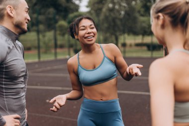 Gülümseyen, heyecanlı Afro-Amerikan kadın şık spor giysiler giyiyor, dışarıda antrenman yaptıktan sonra arkadaşlarıyla konuşuyor. Motivasyon, spor konsepti
