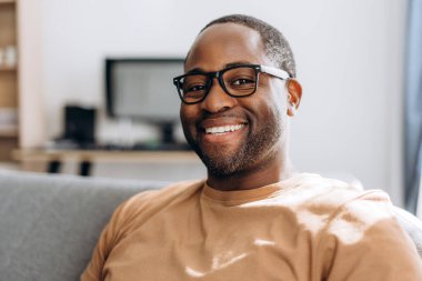 Gözlüklü, gülümseyen Afro-Amerikan adam dairesindeki koltukta oturan kameraya bakıyor.