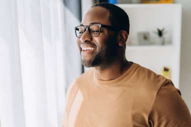 Gözlüklü, gülümseyen Afro-Amerikan adam pencereden dışarı bakarak dairesine poz veriyor.