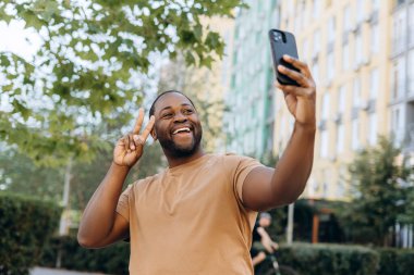 Gülümseyen Afrikalı Afro-Amerikalı bir adamın portresi. V işareti dışarıda telefonda selfie çekiyor.