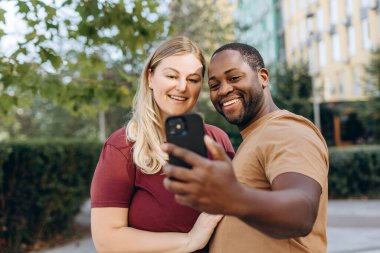 Gülümseyen romantik çift cep telefonuyla selfie çekiyor, şehirde poz veriyor, sarılıyorlar.