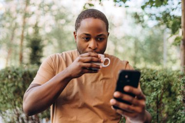 Genç Afrikalı Amerikalı bir adam kafede kahve içiyor, cep telefonu kullanıyor, mesaj okuyor.