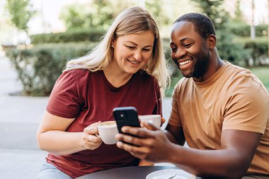 Afro-Amerikalı mutlu bir adam ve kahve fincanlarıyla açık kafede telefon kullanan beyaz bir kadın.