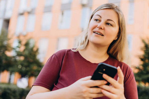 Portrait of beautiful plus size Caucasian woman posing with smartphone looking to the side in the city