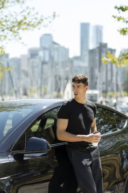 Genç işadamı elinde kahve tutuyor ve şehir merkezinde elektrikli arabasına yaslanıyor.