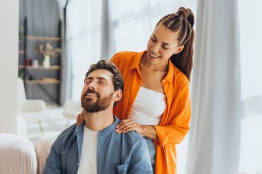 Gülümseyen mutlu arkadaşların portresi, genç güzel bir kadın, sakallı adama boyun masajı yaparken evdeki rahat koltukta oturuyor. Sağlık hizmeti kavramı