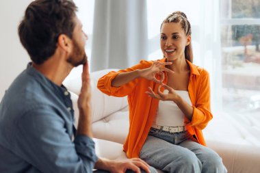 Pozitif sakallı adam portresi ve çekici gülümseyen Latin kadın, çevirmen, evdeki rahat kanepede oturmuş işaret diliyle konuşuyor. İletişim konsepti