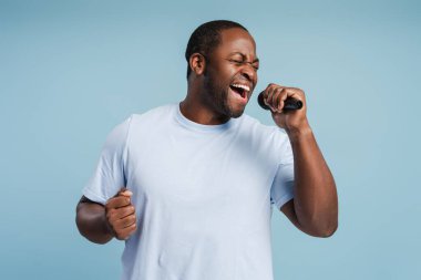 Genç Afrikalı Amerikalı bir adamın portresi. Mikrofonla duygusal şarkı söylüyor. Stüdyoda mavi arka planda izole edilmiş.