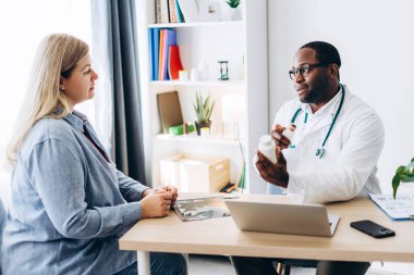 Beyaz önlüklü Afro-Amerikalı erkek doktor klinikteki kadın için ilaç şişesi gösteriyor. Sağlık, tıp konsepti