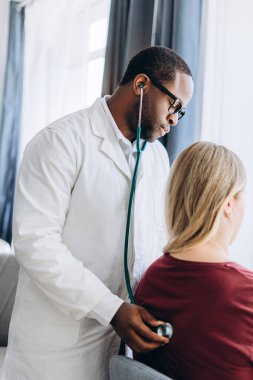 Steteskopla klinikteki kadın kalp atışlarını dinleyen Afrikalı Amerikalı erkek doktor. Sağlık ve kardiyoloji kavramı