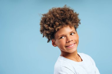 Gülümseyen, neşeli Afro-Amerikalı bir çocuğun portresi, kıvırcık saçlı, başka yöne bakan bir genç, mavi arka planda izole edilmiş bir model, kopyalama alanı. Reklam kavramı