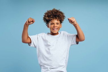 Güçlü, gülümseyen Afrikalı Amerikalı bir gencin portresi, pazılarla el ele tutuşan, kameraya bakan duygusal bir çocuk. Mavi arka planda izole edilmiş bir model. Reklam kavramı