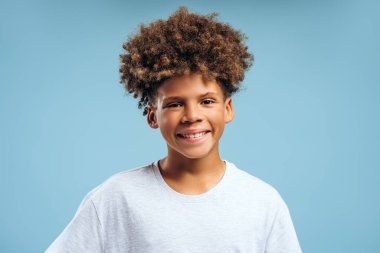 Mutlu, gülümseyen Afrikalı Amerikalı bir gencin portresi, kameraya bakan kıvırcık saçlı bir çocuk. Mavi arka planda izole edilmiş küçük bir model. Reklam, diş bakımı konsepti