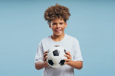 Gülümseyen, mutlu, çekici çocuk, kıvırcık saçlı çocuk futbol topuna bakıyor kameraya bakıyor, mavi arka planda izole edilmiş poz veriyor, reklam kavramı.