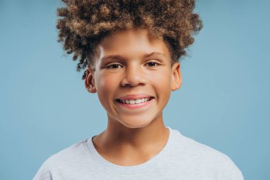 Yakın plan, gülümseyen, kıvırcık saçlı mutlu Afro-Amerikan çocuk portresi, mavi arka planda izole edilmiş kameraya bakan genç. Reklam, diş konsepti