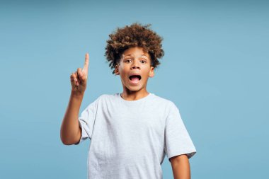 Şok olmuş, kıvırcık saçlı, ağzı açık, kameraya bakan ve mavi arka planda izole edilmiş bir fikri olan Afro-Amerikan çocuğun portresi. Reklam kavramı