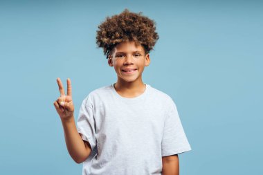 Mutlu, çekici, kıvırcık saçlı beyaz tişört giyen, barış işareti gösteren, mavi arka planda izole edilmiş poz veren Afro-Amerikan çocuk portresi. Zafer konsepti