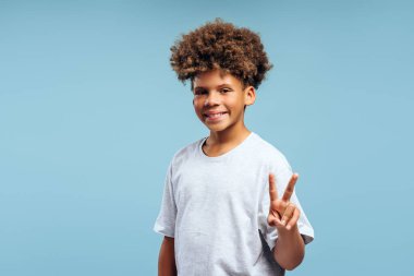 Çekici, gülümseyen, kıvırcık saçlı, barış işareti gösteren, mavi arka planda izole edilmiş, fotokopi çeken Afro-Amerikan çocuk. Hareketli kavram