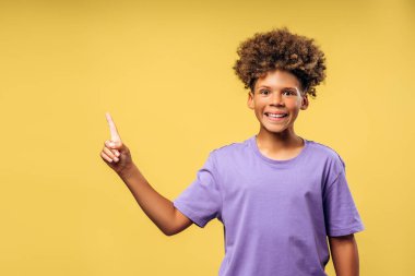 Mutlu, pozitif, kıvırcık saçlı, fotokopi alanını gösteren ve kameraya bakan Afro-Amerikan çocuk portresi. Sarı arka planda izole edilmiş bir model. Reklam, alışveriş konsepti