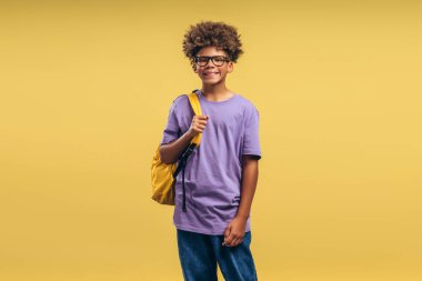 Zeki, gülümseyen, mutlu Afro-Amerikan çocuk, kıvırcık saçlı, sırt çantalı, şık gözlüklü öğrenci, sarı arka planda tek başına duran kameraya bakıyor. Eğitim kavramı
