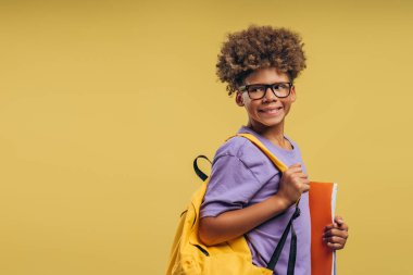 Çekici, gülümseyen Afrikalı Amerikalı çocuk, sırt çantası taşıyan öğrenci ve şık gözlük takan kitaplar, başka tarafa bakıyor, sarı arka planda izole edilmiş poz veriyor. Eğitim kavramı