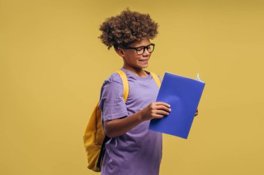 Mutlu, gülümseyen Afrikalı Amerikalı bir çocuğun portresi, şık gözlük takan bir öğrenci, sırt çantası tutuyor ve sarı arka planda izole edilmiş kitap okuyor. Eğitim kavramı
