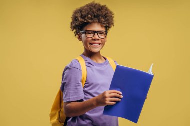 Gülümseyen Afrikalı Amerikalı bir çocuğun portresi, şık gözlük takan bir öğrenci, sırt çantası tutuyor ve sarı arka planda izole edilmiş kameraya bakıyor. Eğitim kavramı