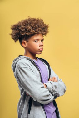 Dalgın, kıvırcık saçlı, kollarını kavuşturmuş, sarı arka planda izole bir şekilde duran kameraya bakan Afro-Amerikan çocuğun portresi. Reklam kavramı