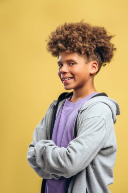 Neşeli, mutlu Afro-Amerikan çocuk, kıvırcık saçlı, çapraz kollu, sarı arka planda yalnız poz veren genç. Reklam kavramı