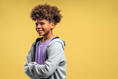 Gülümseyen, mutlu Afro-Amerikan çocuk, kıvırcık saçlı, çapraz kollu genç, sarı arka plan fotokopi alanında yalnız poz veriyor. Reklam kavramı