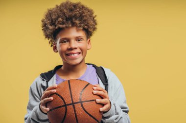 Pozitif Afro-Amerikan çocuk portresi, kıvırcık saçlı basketbol topu tutan, kameraya bakan, sarı arka planda izole edilmiş poz veren. Oyun, spor konsepti