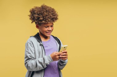 Mutlu, gülümseyen, kıvırcık saçlı, şık kapüşonlu, cep telefonu kullanan, mobil uygulama kullanan, sarı arka planda izole edilmiş, çekici Afrikalı Amerikalı çocuk. Çevrimiçi teknoloji kavramı