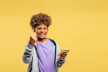 Mutlu Afro-Amerikan çocuk portresi şık saç stiline sahip, cep telefonu kullanan, mobil uygulama kullanan, kazanan, kameraya bakan, sarı arka planda izole edilmiş poz veren. Zafer konsepti