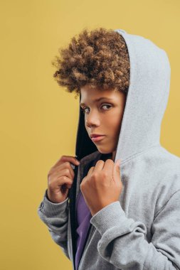 Kafasında kıvırcık saç stili kapüşonlu yakışıklı Afro-Amerikan çocuğun portresi sarı arka planda izole edilmiş bir şekilde kameraya bakıyor. Reklam kavramı