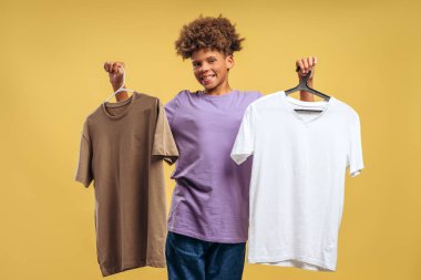 Gülümseyen, çekici Afro-Amerikan çocuk elinde iki tişört, beyaz ve mor, sarı arka planda tek başına duran kameraya bakıyor. Alışveriş konsepti
