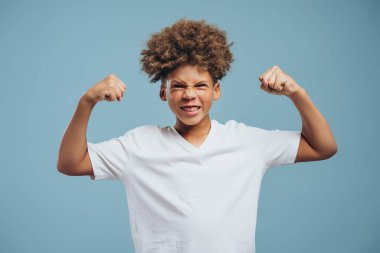 Güçlü, gülümseyen, kıvırcık saçlı, elinde pazıları olan, kameraya bakan, mavi arka planda izole edilmiş poz veren Afrikalı Amerikalı bir çocuğun portresi..